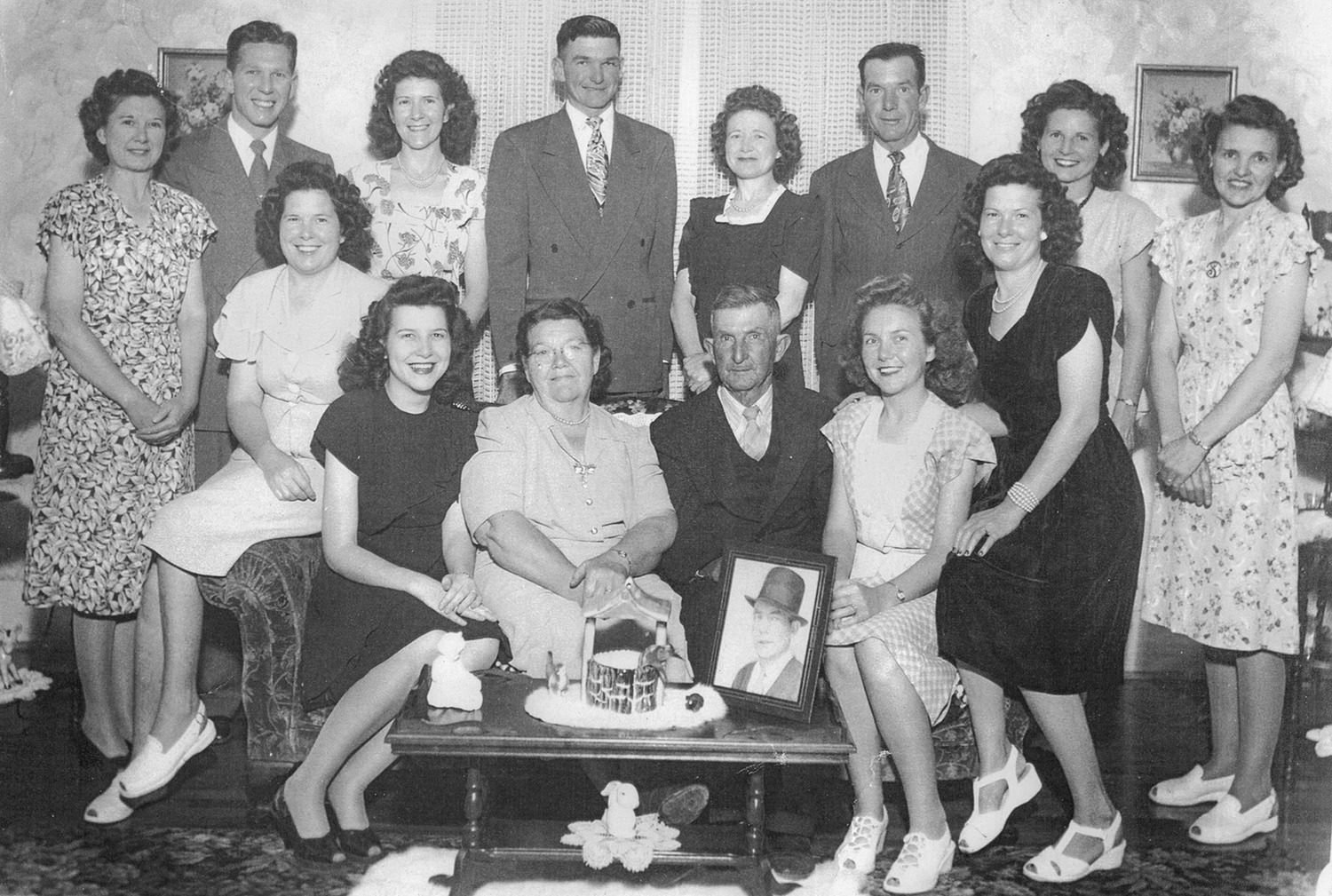 Group of adults and teens posing for a vintage family portrait in a living room, with a framed photo on the coffee table and a cake centerpiece in front of them.