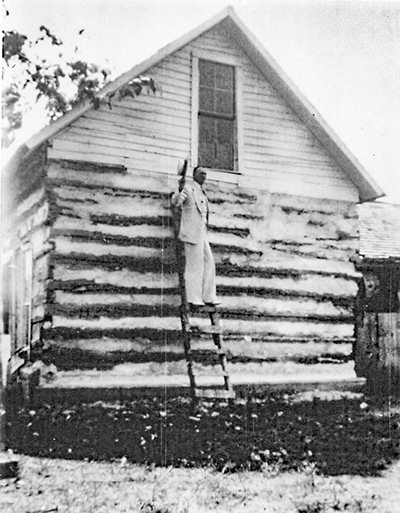 Man on a tall ladder leaning against a white wooden log cabin, holding a long pole.