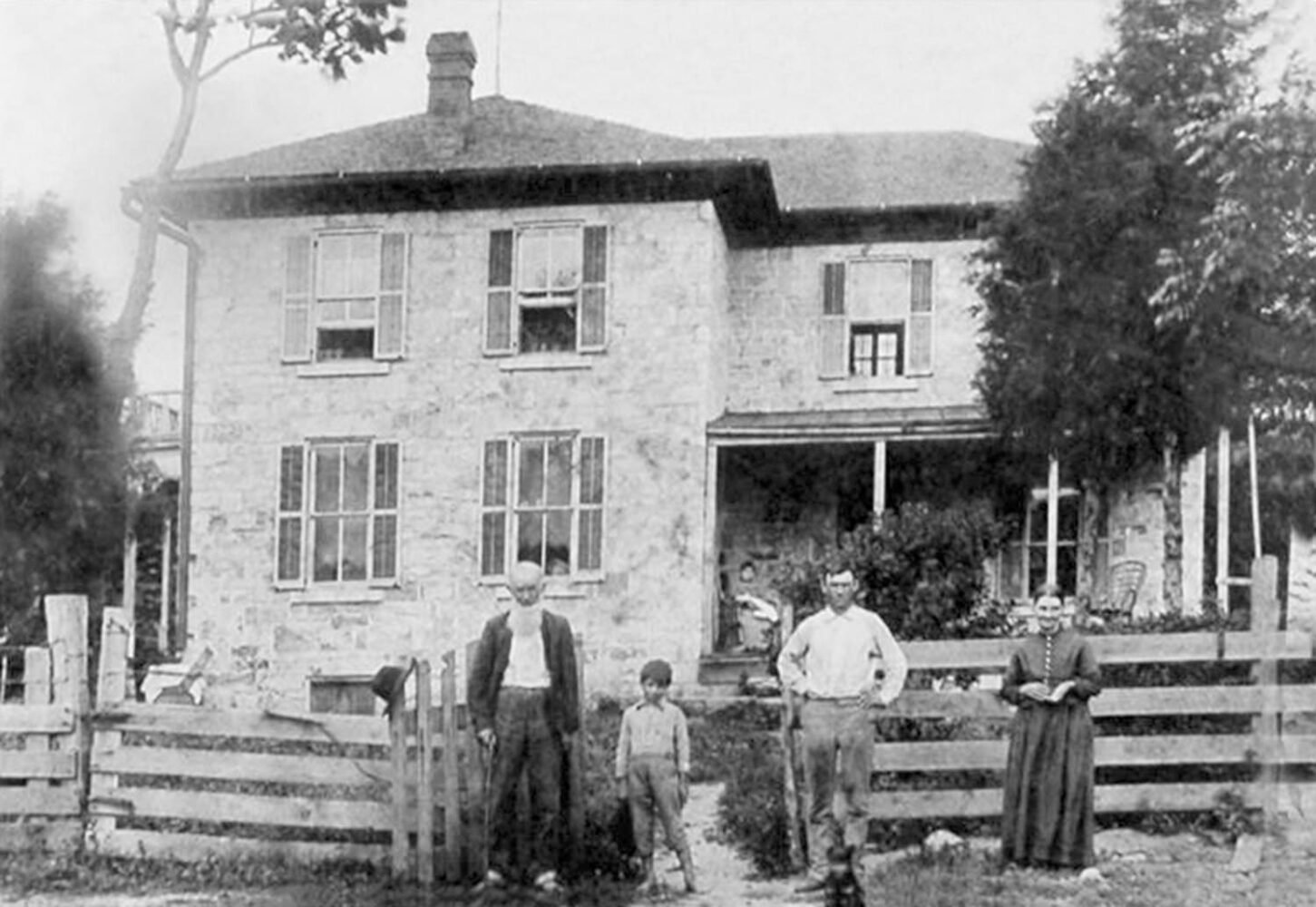 Stone two-story house with shuttered windows and a wraparound porch; four people—two men, a boy, and a woman—stand at a wooden fence in front.