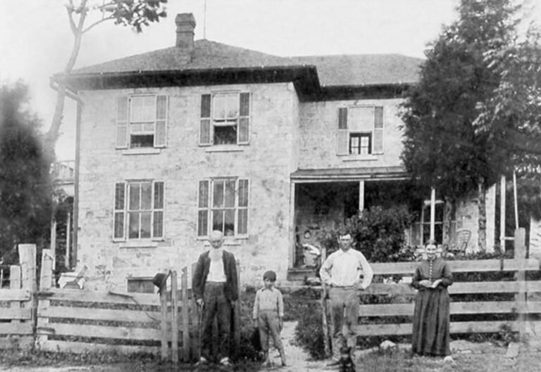 Stone two-story house with shuttered windows and a wraparound porch; four people—two men, a boy, and a woman—stand at a wooden fence in front.