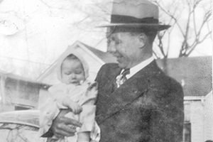 Man in a dark suit and hat holding a baby wrapped in a blanket outdoors, with a house and bare trees in the background.