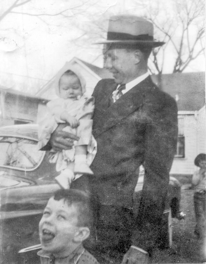 Man in a suit and fedora holding a baby, smiling outdoors with a young boy in front; vintage car and houses in the background.