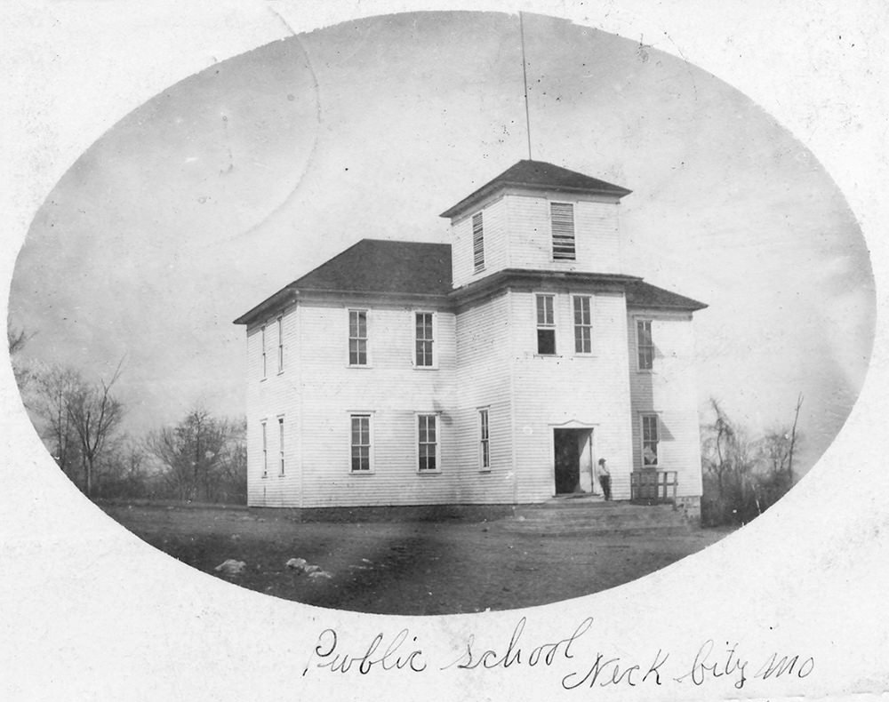 Historic two-story wooden public schoolhouse with a central tower, photographed in an open yard (vintage image).