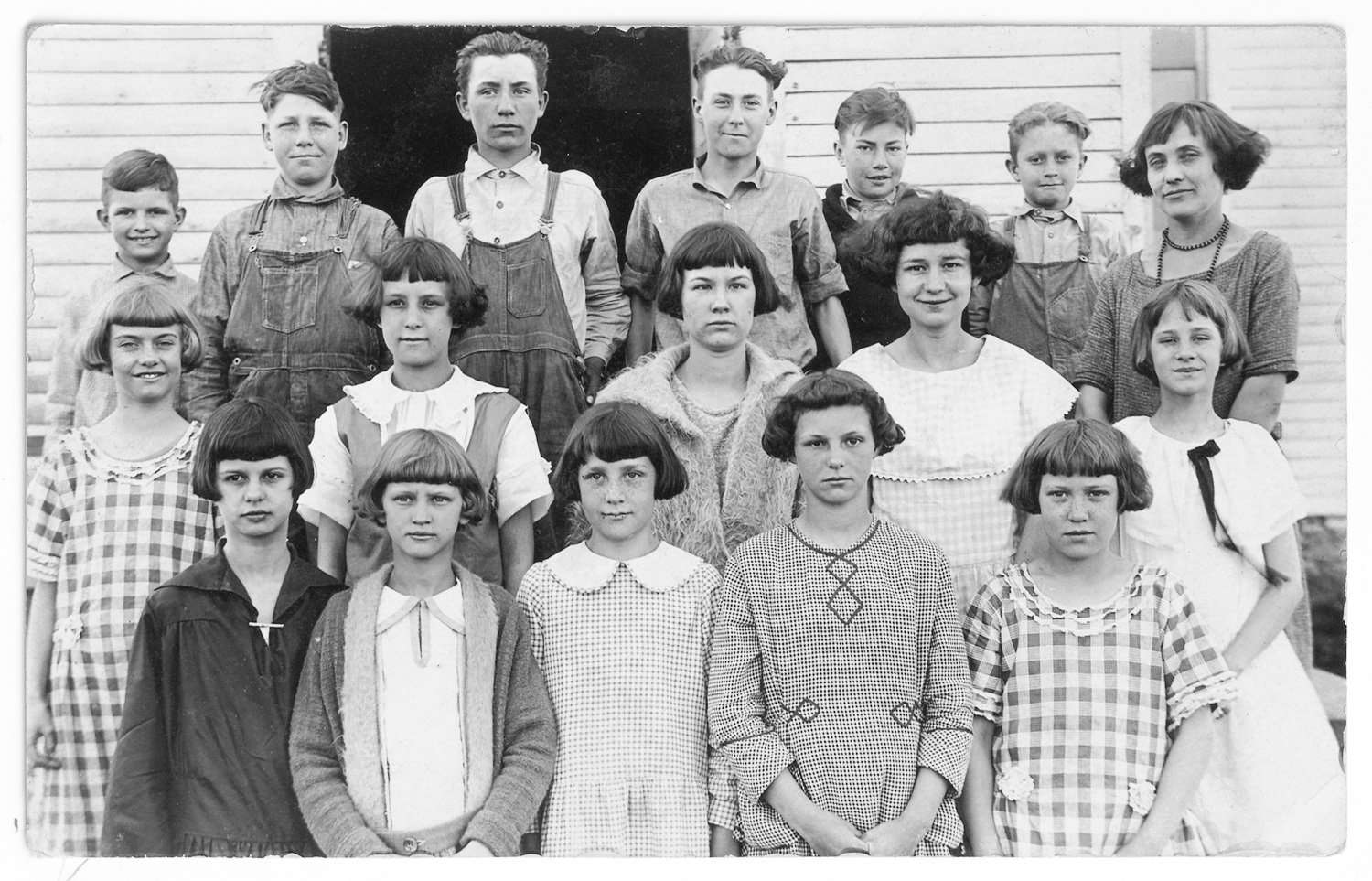 Group portrait of a class: 15 children and a female teacher posing outside a wooden building, arranged in rows.