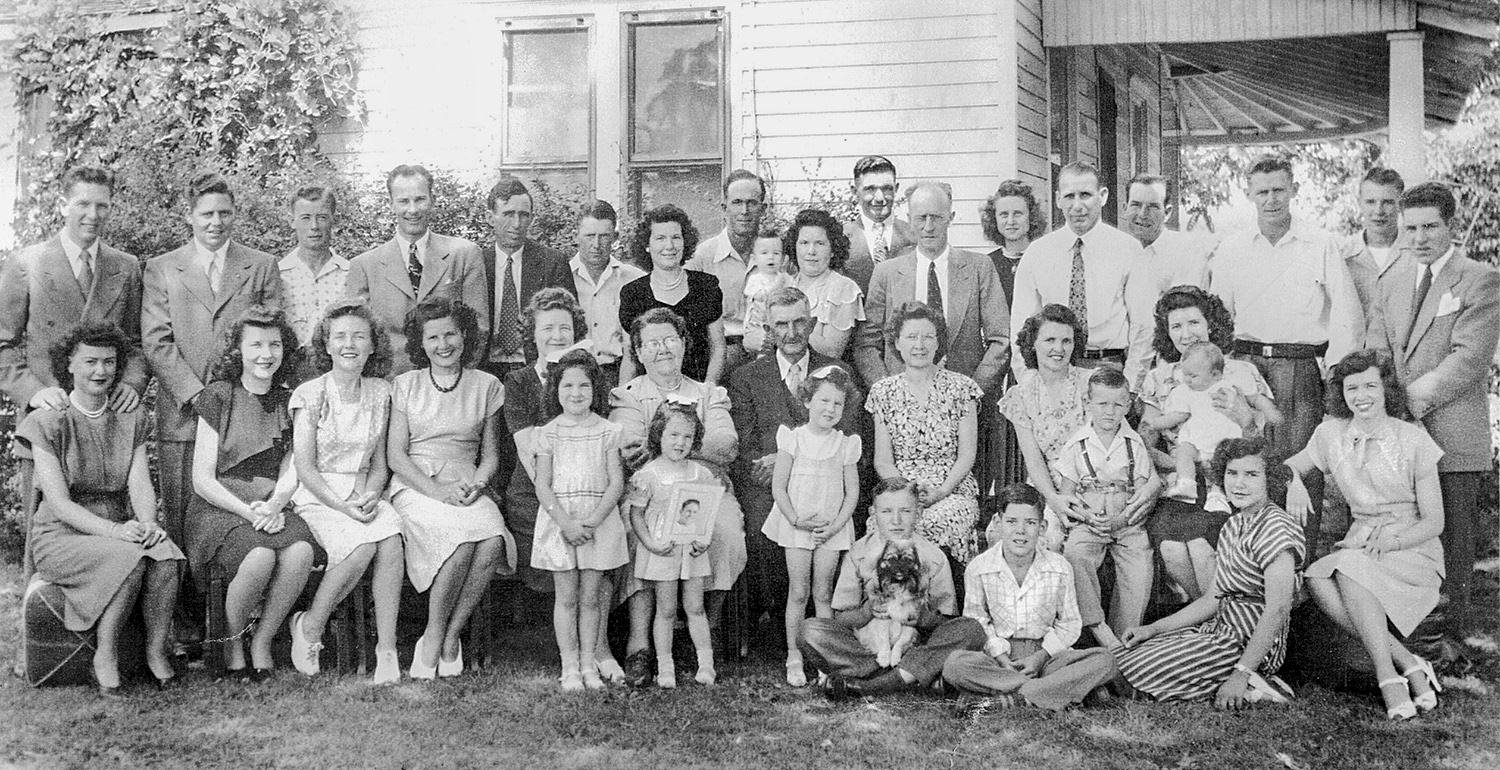 Large family group posing for a formal portrait on the front lawn, with dozens of adults and children in spring clothing in front of a house.
