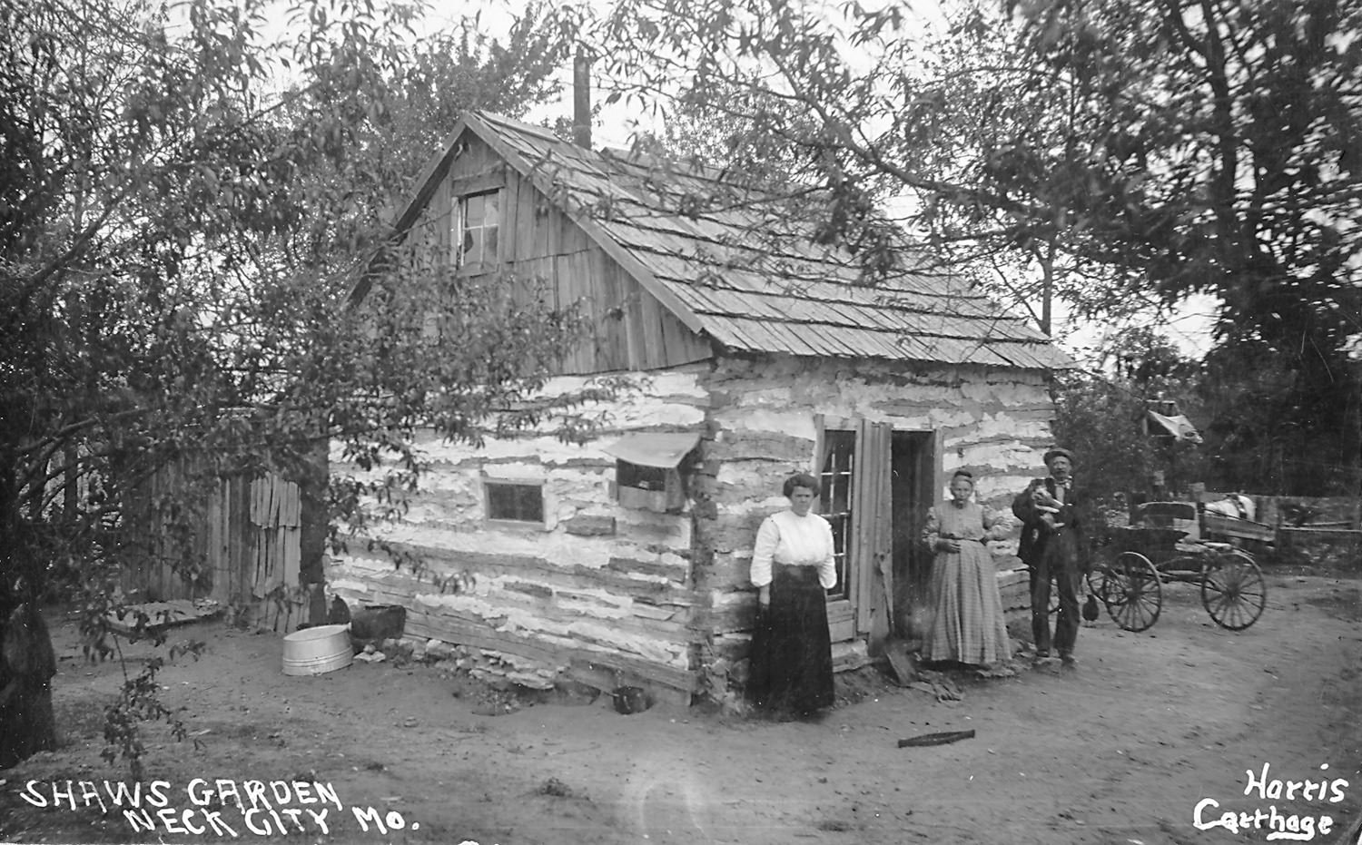 Historic rural scene: a rough log cabin with a wooden shingled roof, three people in period dress standing by the door, and a wagon in the yard.