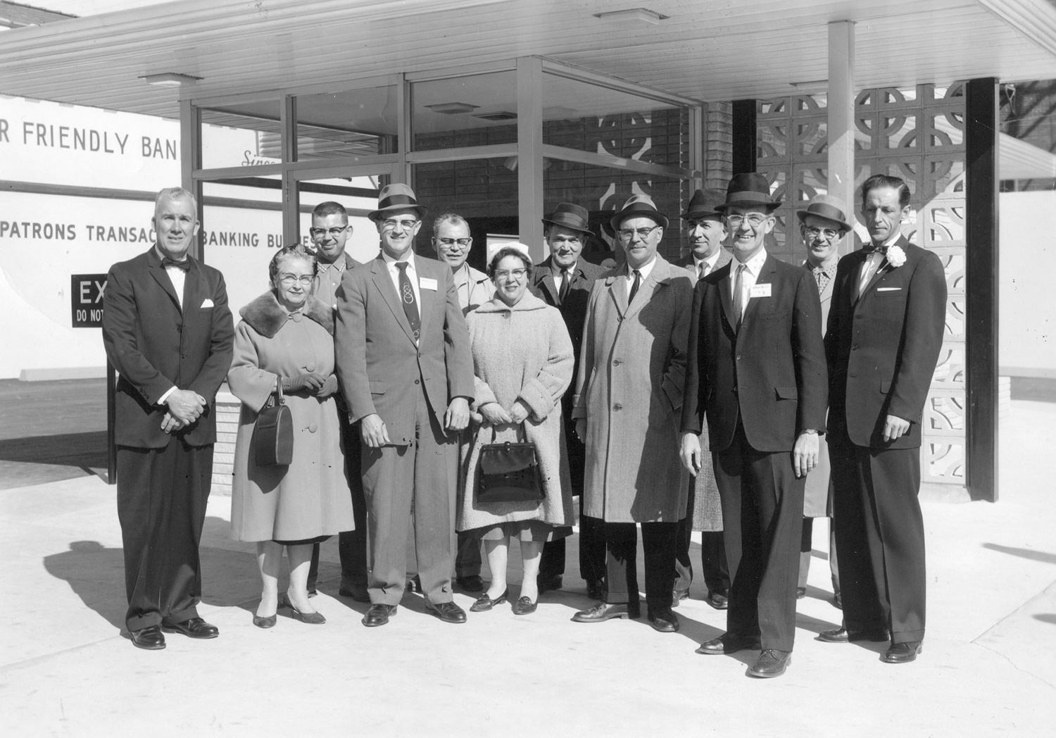 Large group of formally dressed men and two women posing outside a mid‑century bank building for a group photo.