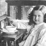 Young girl with wavy hair sits at a wooden table, reaching for a teacup on a saucer in a home setting.