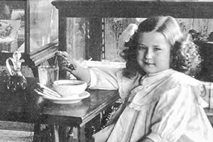 Young girl with wavy hair sits at a wooden table, reaching for a teacup on a saucer in a home setting.