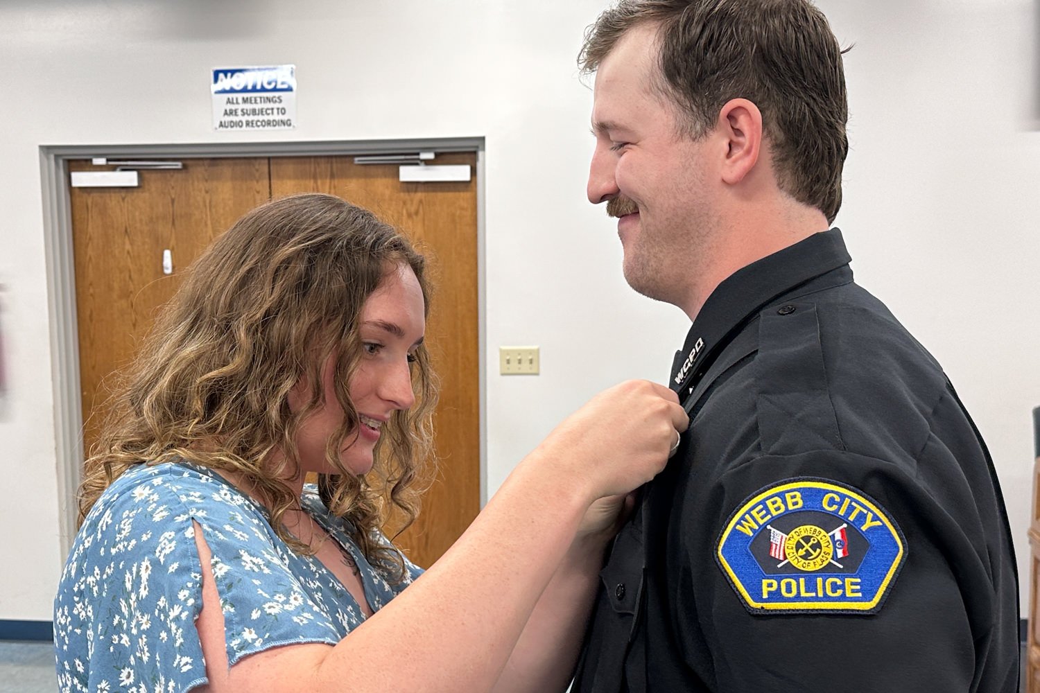 Woman pins the lapel of a Webb City Police officer's uniform, both smiling in a hallway.
