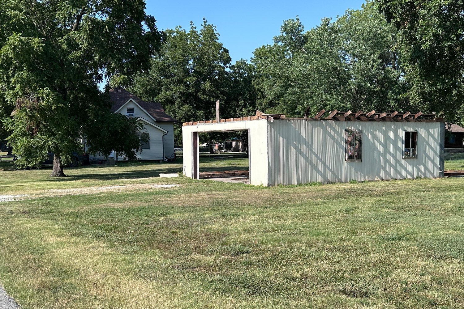 White unfinished building with an open doorway and exposed roof beams beside a house in a grassy park under a blue sky.
