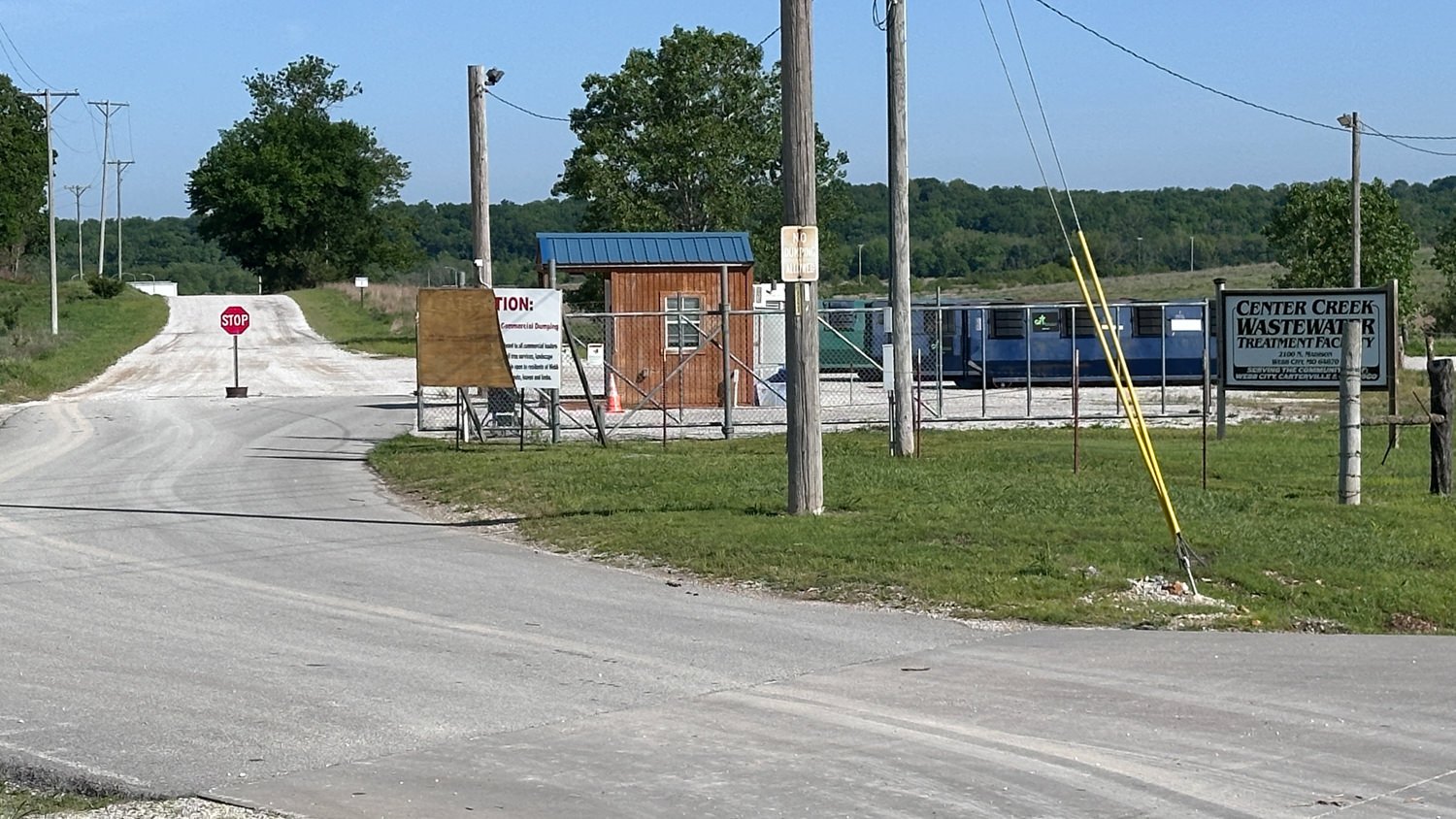 Rural road leads to a fenced wastewater treatment facility with a small wooden office, a stop sign, power lines, and a sign reading Center Creek Wastewater Treatment Facility.