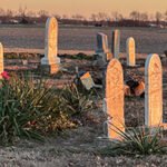Desert cemetery at dusk with tall stone gravestones and desert shrubs in the foreground.
