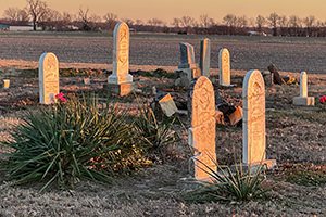 Desert cemetery at dusk with tall stone gravestones and desert shrubs in the foreground.