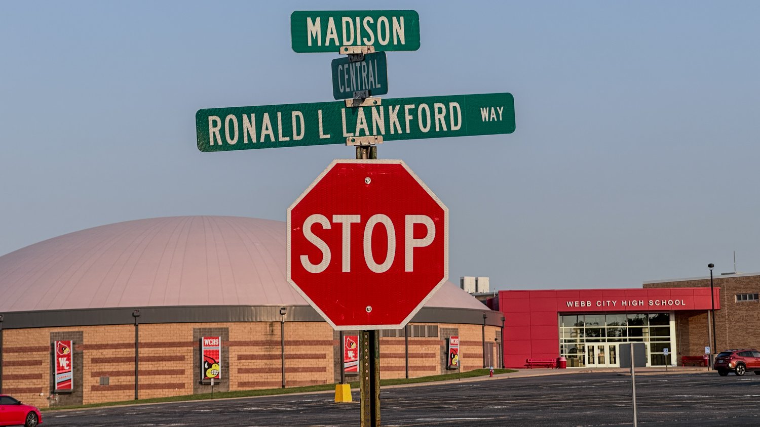 Stop sign at the intersection with Madison Central and Ronald L. Lankford Way in front of a high school campus, with a pink-domed building nearby.