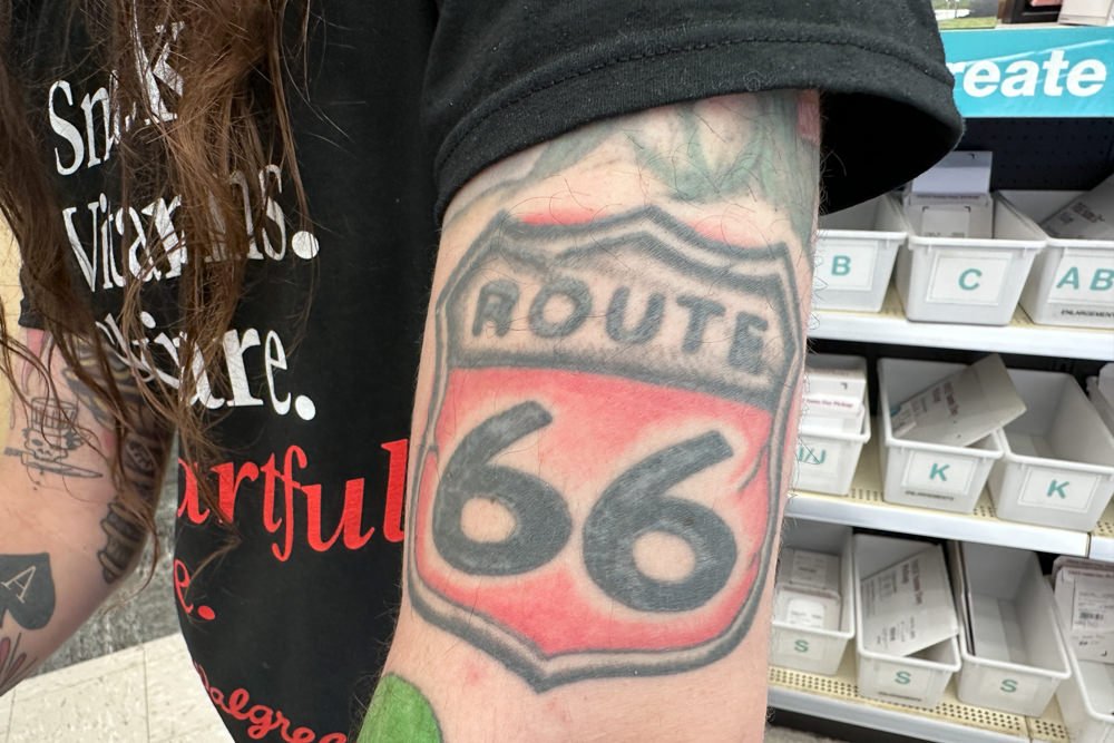 Forearm tattoo of Route 66 shield in red and gray, with store shelves and labeled bins in the background.