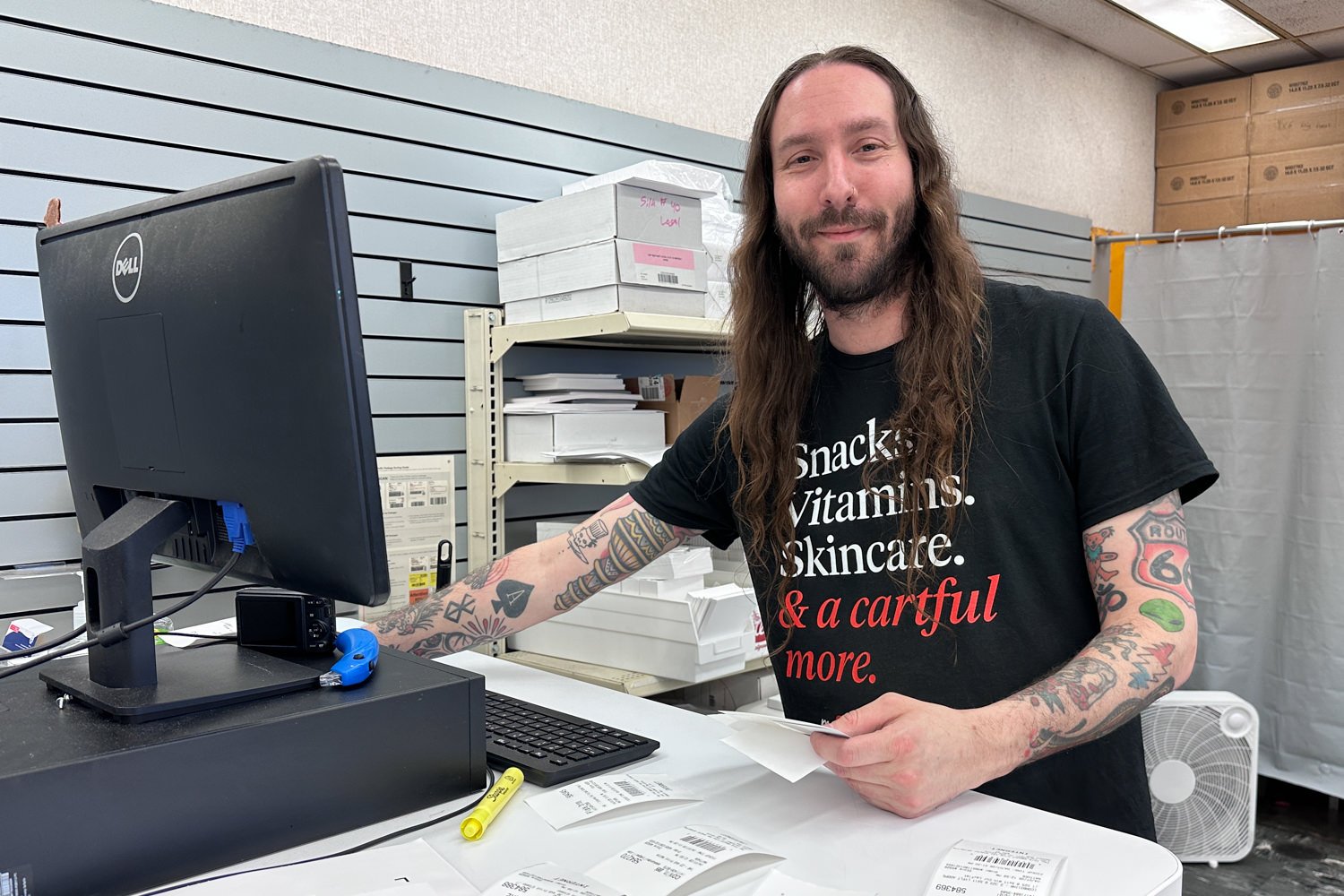 Cashier with long hair and tattoos at a checkout counter, handling receipts near a Dell computer in a store.html description of the setting