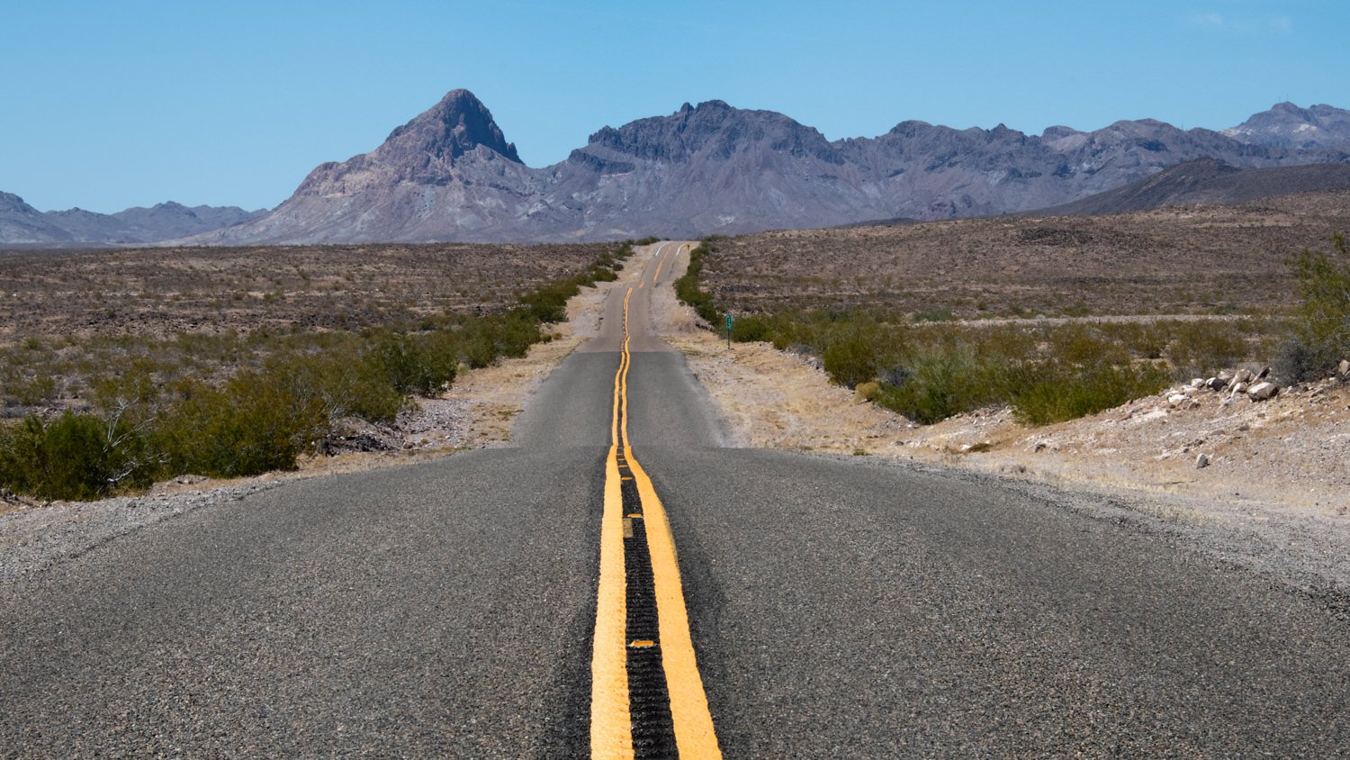 Desert highway stretching toward distant rugged mountains under a clear blue sky, with a double yellow line down the center and sparse shrubs on each side.