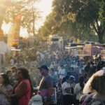 Crowd of festival-goers at an outdoor market with tents, flags, and booths on a sunny day.