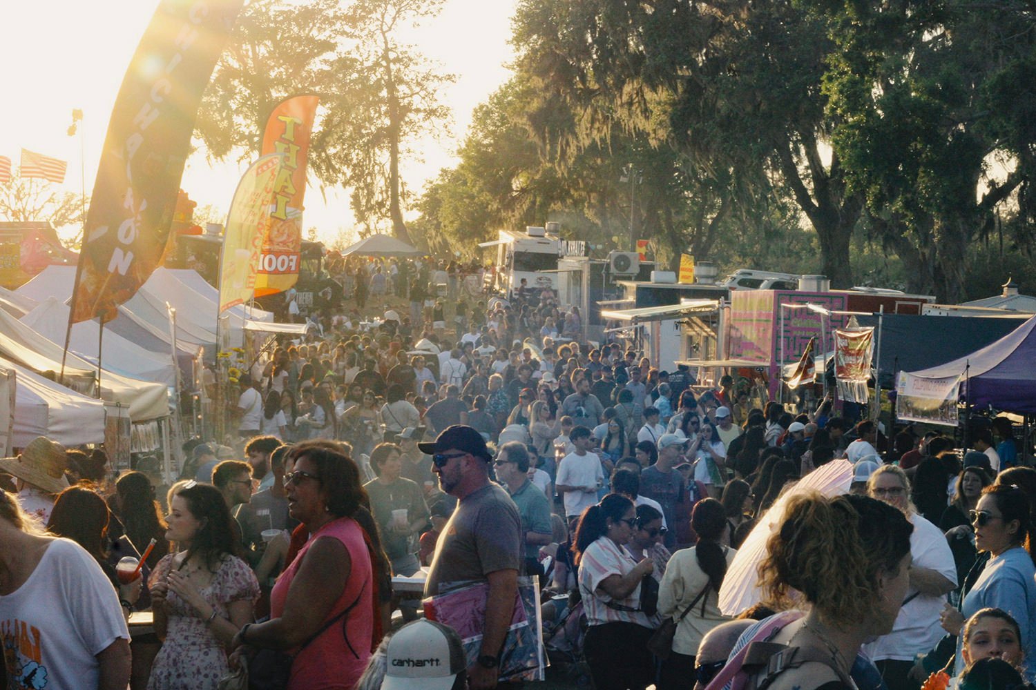 Crowd of festival-goers at an outdoor market with tents, flags, and booths on a sunny day.