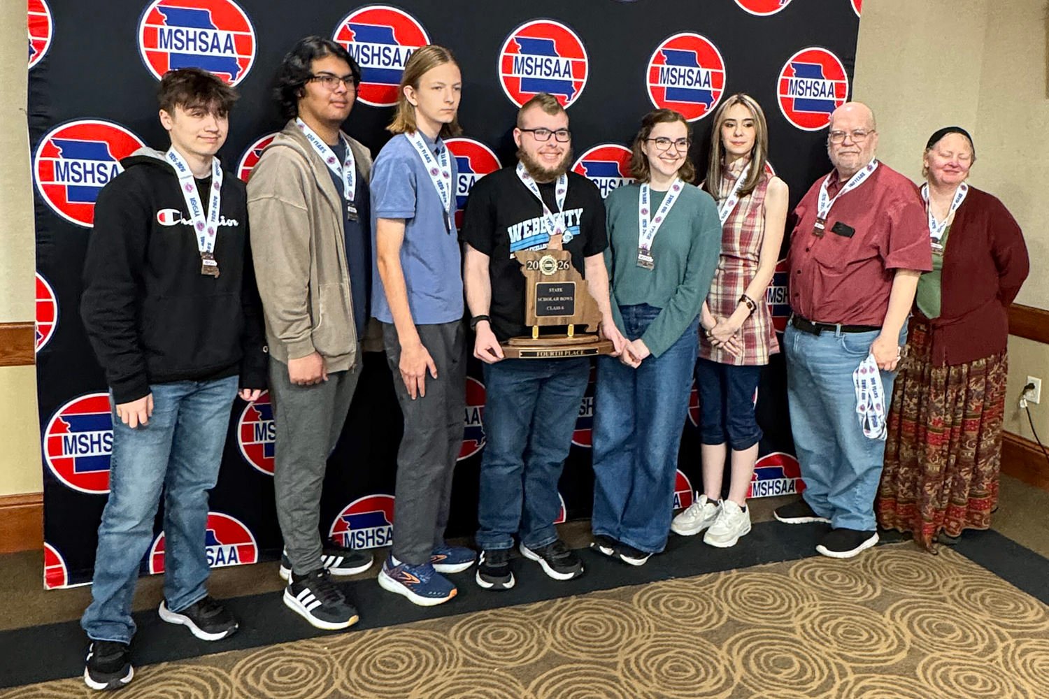 Eight students and two adults pose with a trophy in front of a MSHSAA backdrop, all wearing medals.