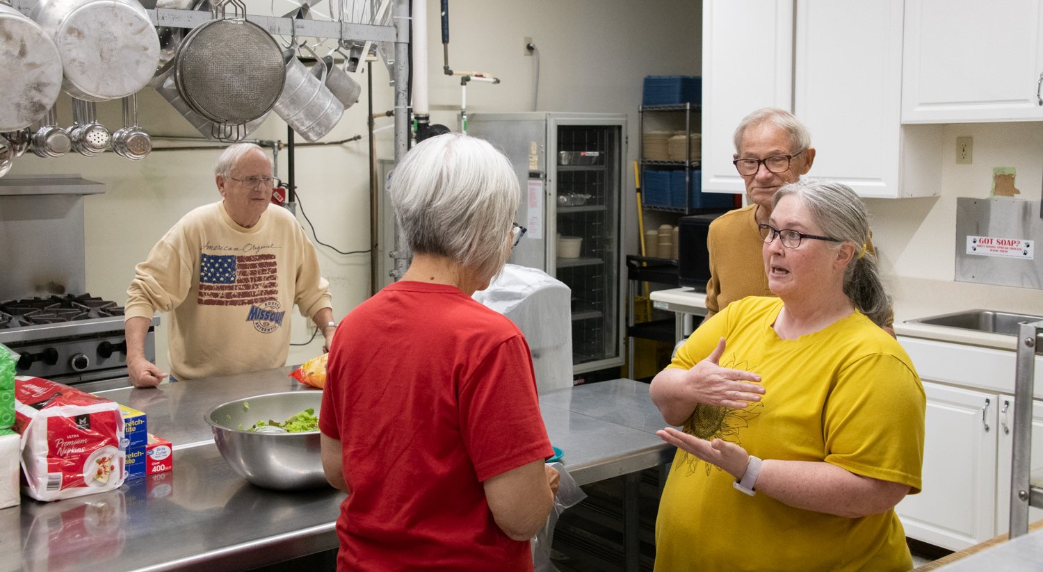 Four older adults stand in a commercial kitchen, chatting near a stainless steel prep table with a bowl of chopped vegetables and bags of supplies nearby.