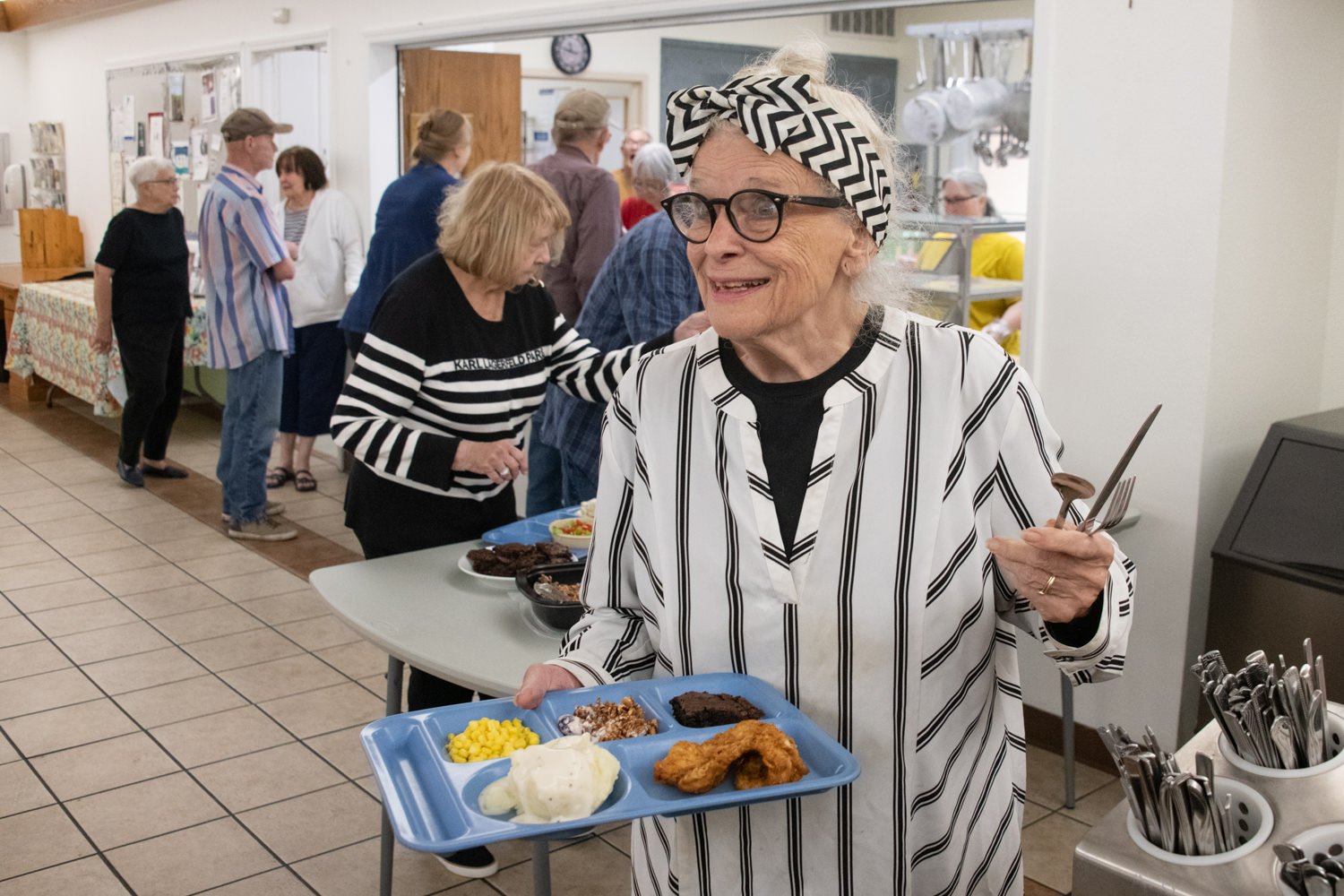 Smiling elderly woman in a black-and-white striped outfit and headband, serving food in a community kitchen, holding a blue tray with corn, mashed potatoes, fried chicken, and dessert pieces.