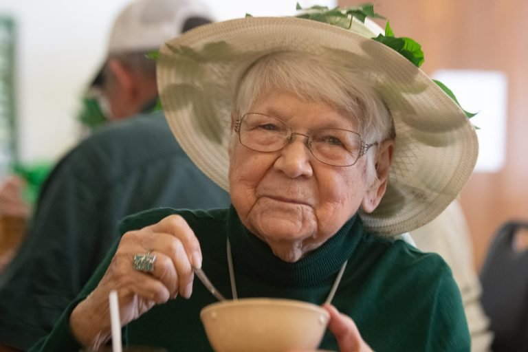 Elderly woman wearing a wide-brimmed hat with green leaves, scooping food from a bowl with a spoon.