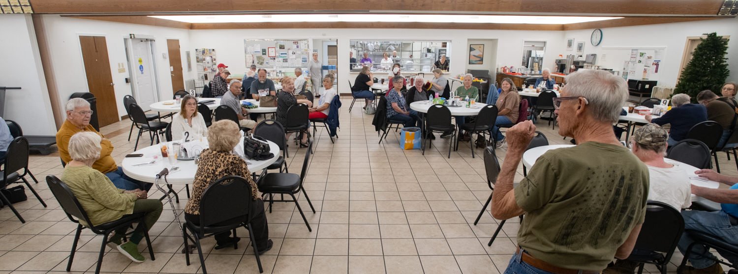 A large room filled with round tables and seniors seated, chatting and eating at a community gathering hall.