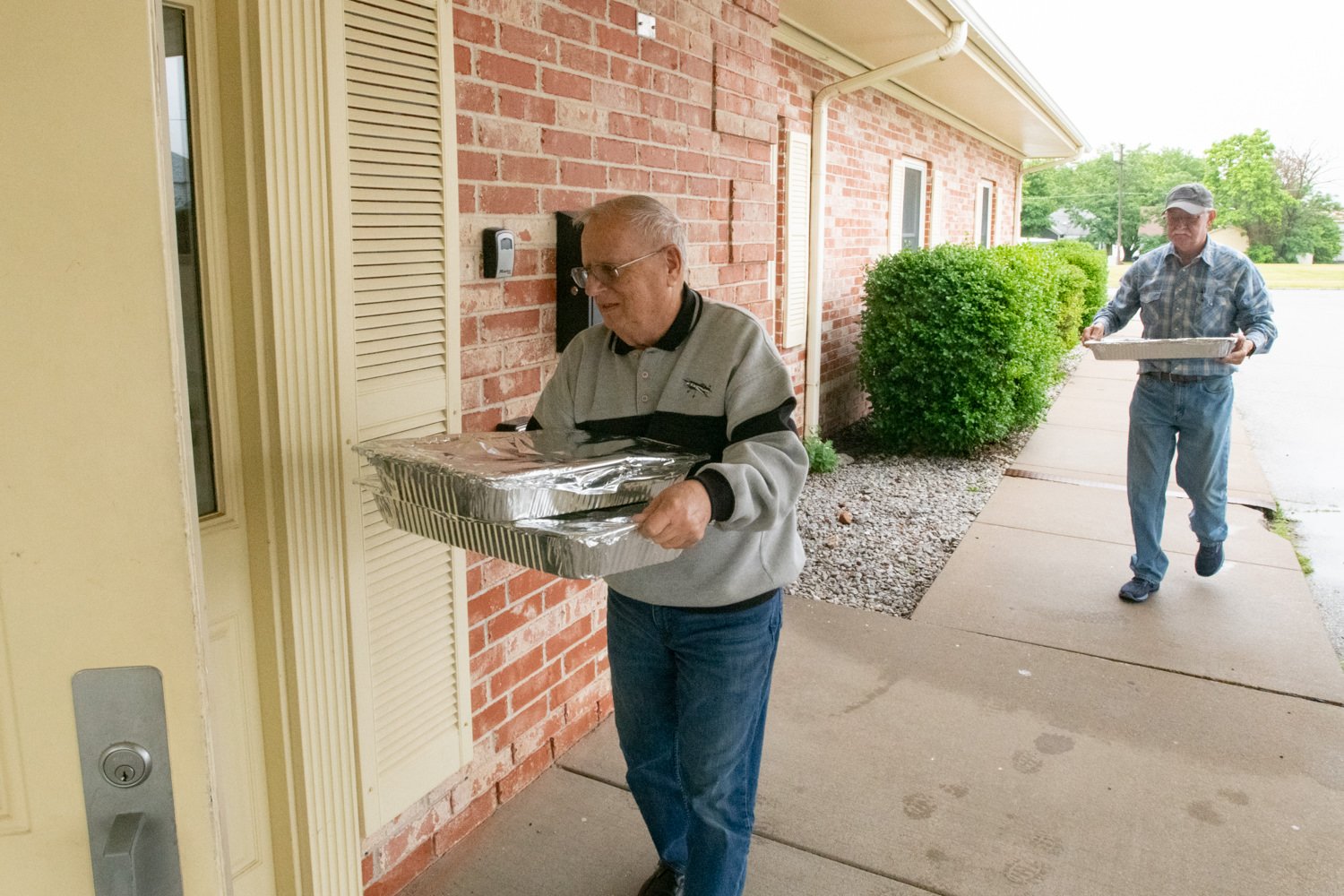 Two elderly men deliver foil-covered food trays to a brick house, one at the door and another approaching with a tray.