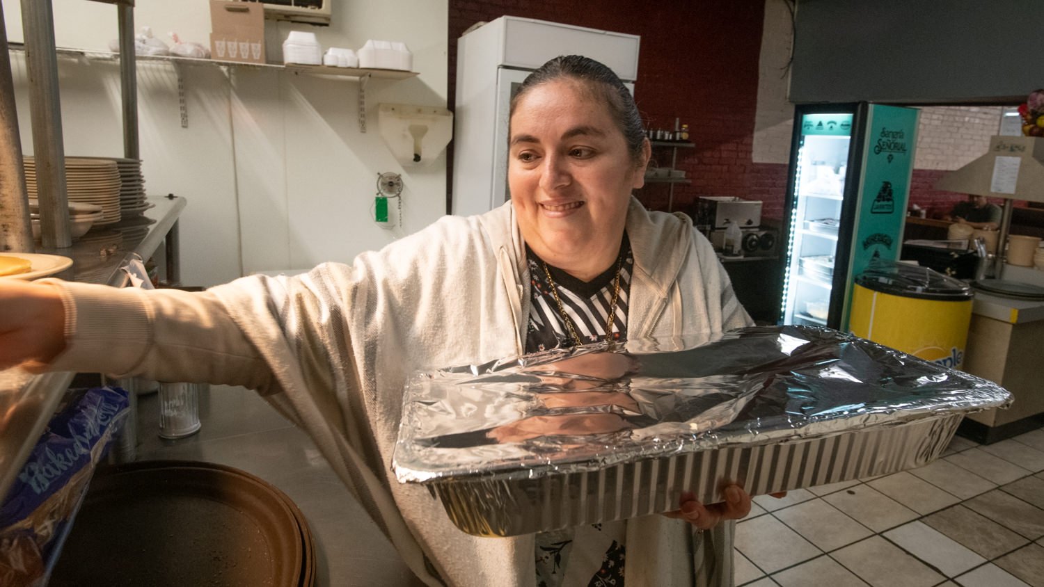 Smiling woman in a kitchen holding a large aluminum foil tray toward the viewer.