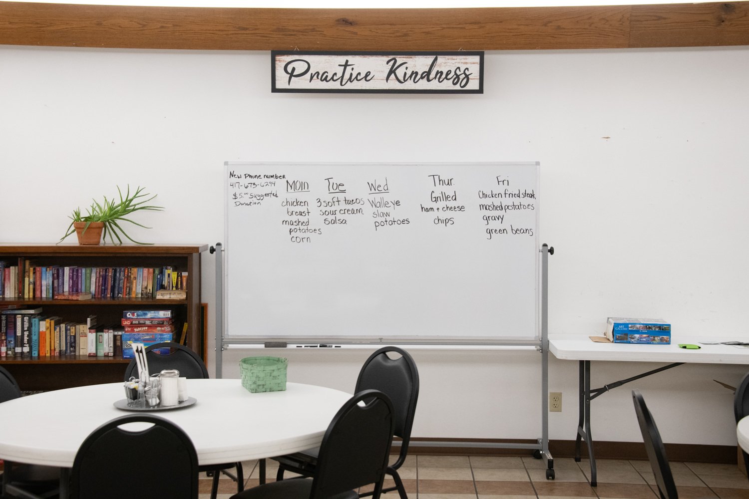 Community room with round tables, black chairs, a bookshelf, a potted plant, and a whiteboard menu under a 'Practice Kindness' sign.