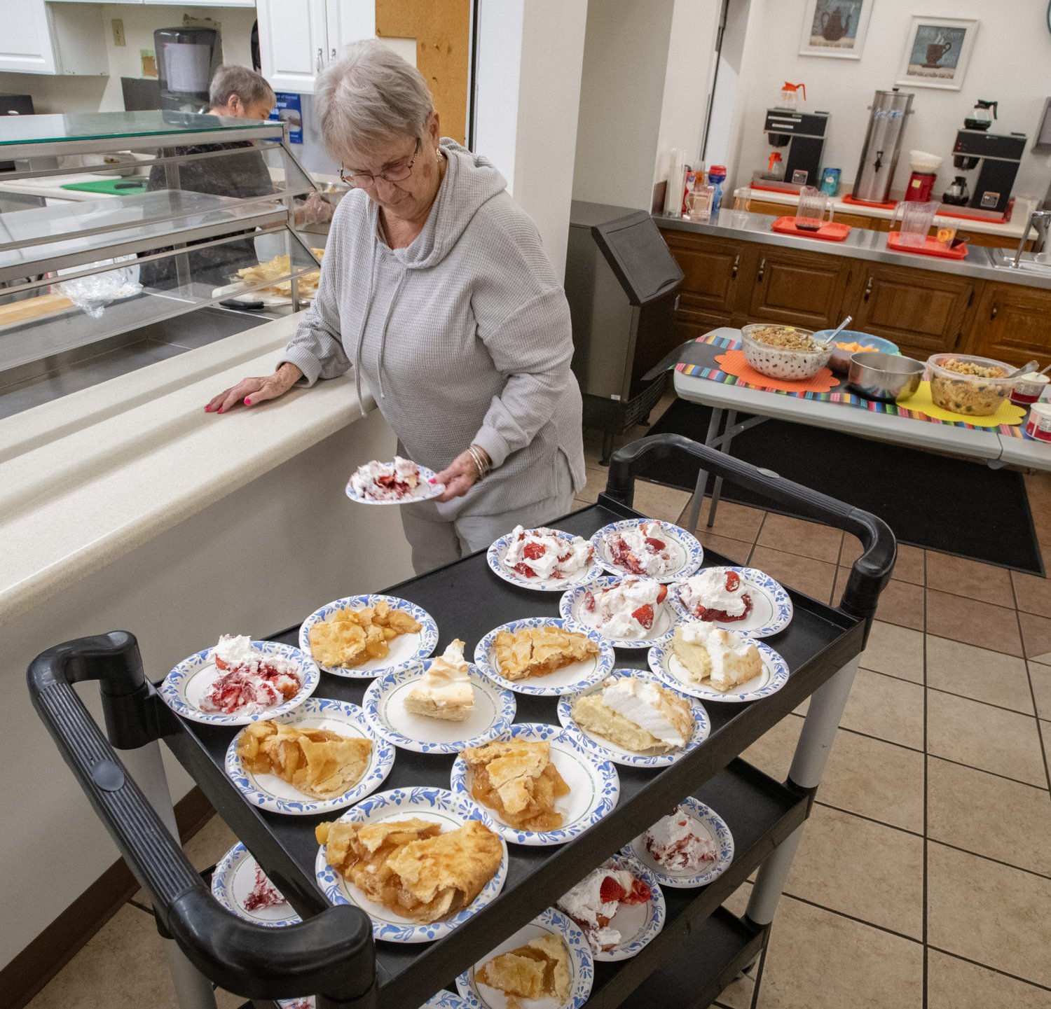 Older woman in a gray hoodie places dessert plates on a rolling cart in a cafeteria, with pies topped with whipped cream and strawberries nearby.