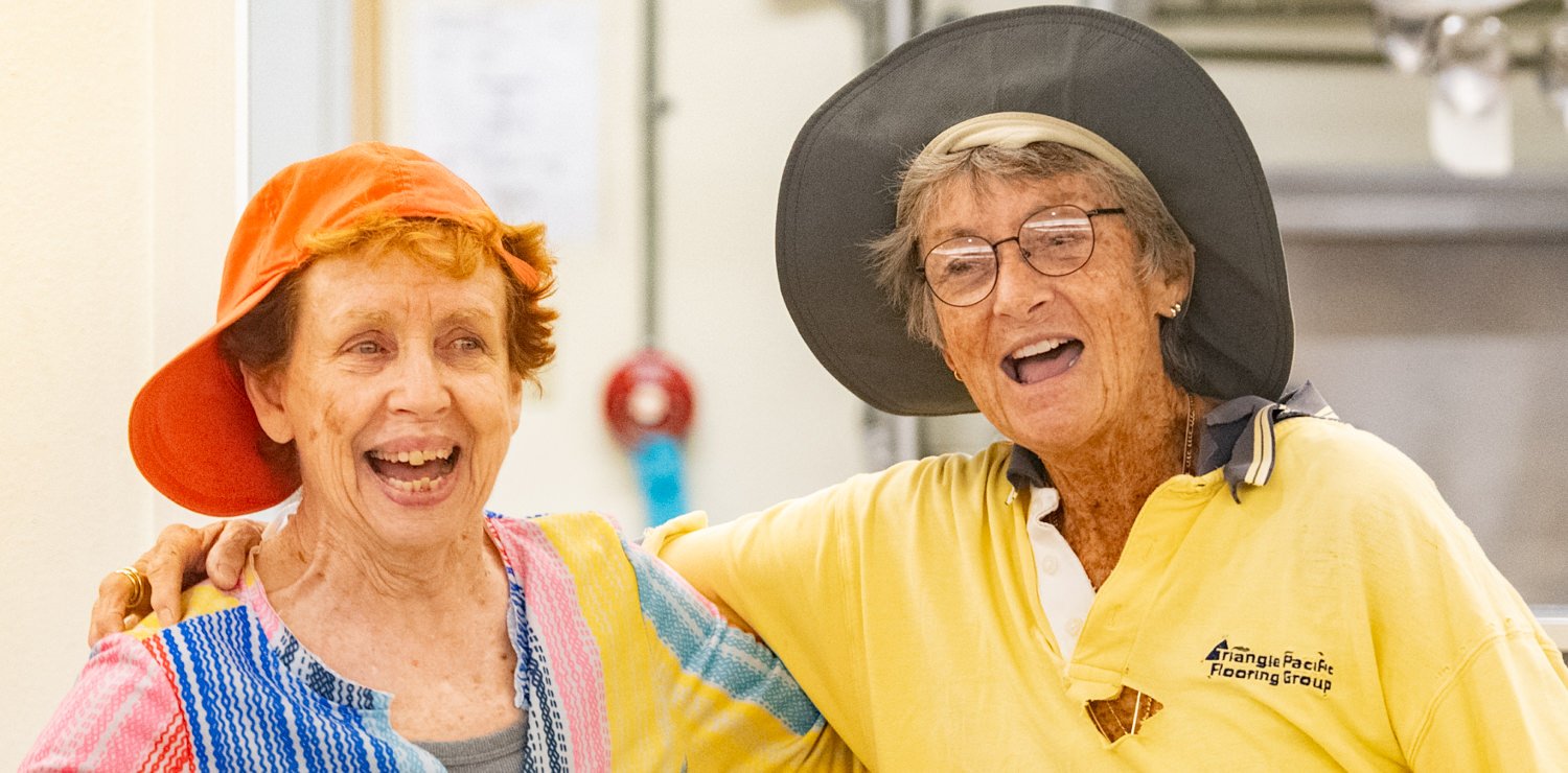 Two elderly women with hats smiling and arms around each other in a workroom.