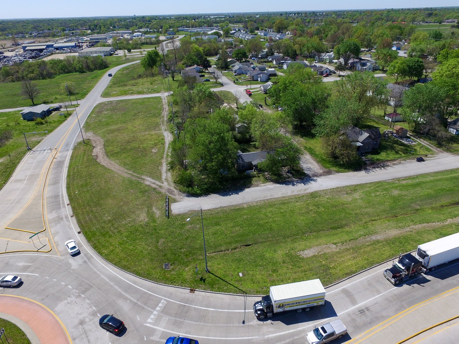 Aerial view of a busy roundabout with trucks and cars, bordered by green fields and a small residential area nearby.
