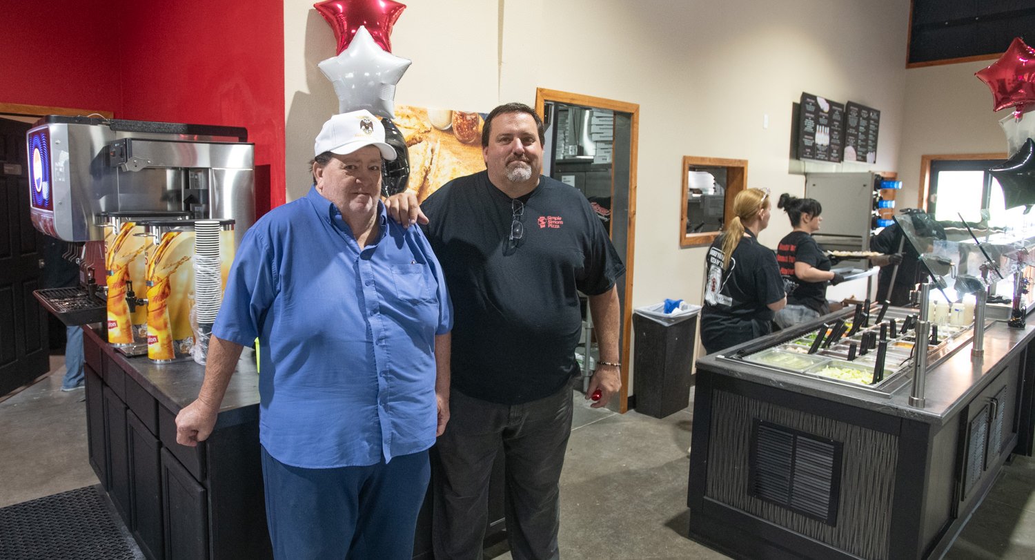 Two men pose for a photo inside a pizza shop, with a toppings counter and staff in the background.