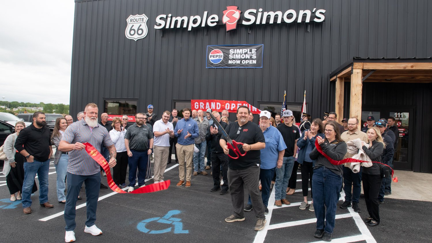 A group of people outdoors at a ribbon-cutting ceremony in front of a black building with Simple Simon's sign and a 'GRAND OPEN' banner behind them.