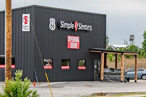 Dark gray Simple Simon's building with an 'OPENING SOON' sign and Route 66 logo, in a parking lot with a wooden overhang.