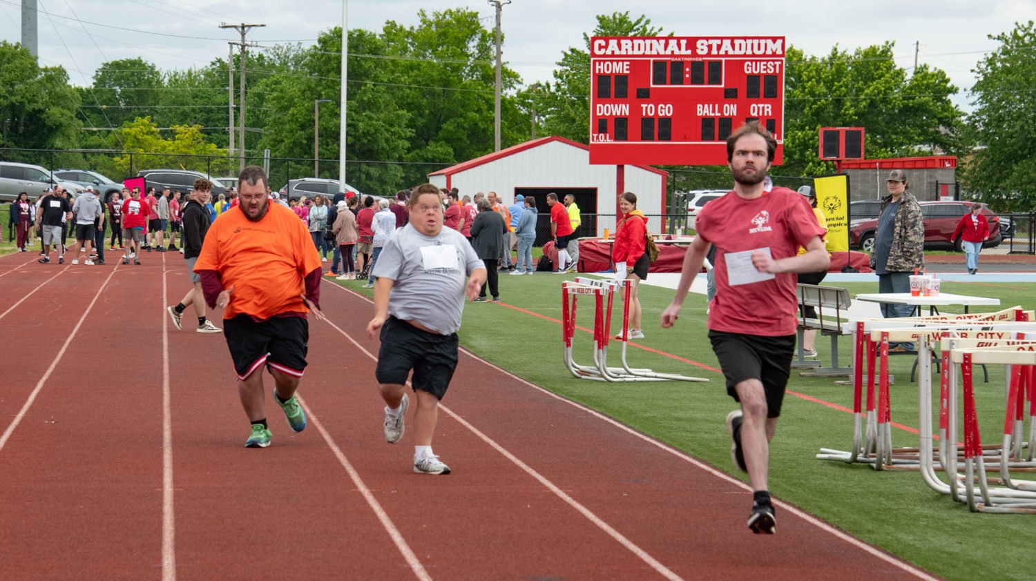 Runners sprint toward the finish on a track with a crowd watching; scoreboard reads Cardinal Stadium in the background.