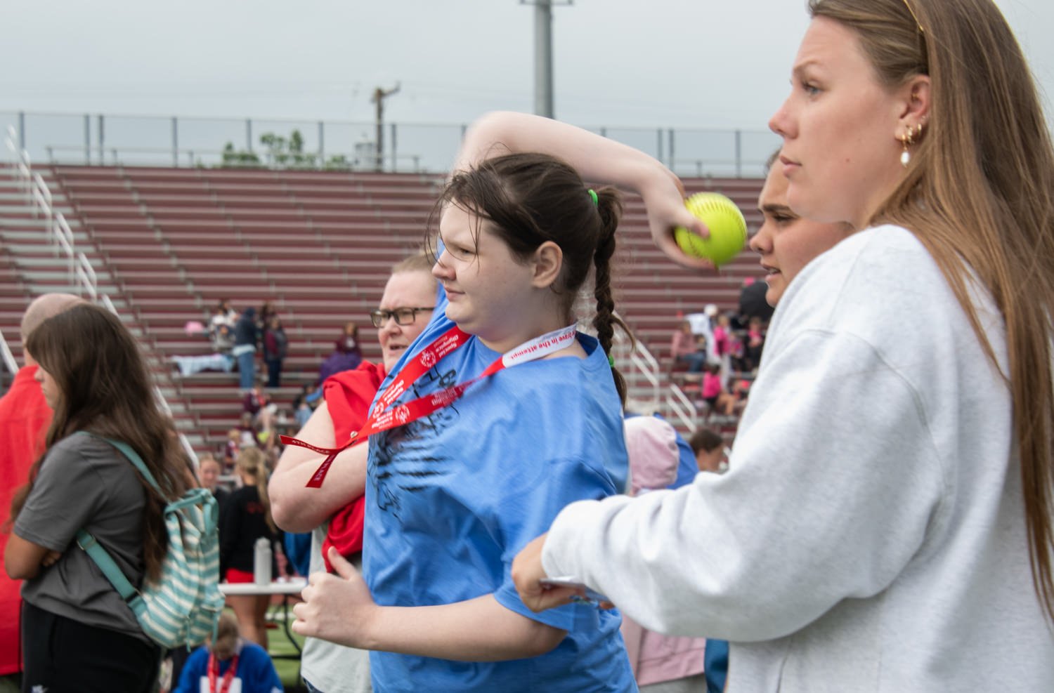 Girl in a blue shirt with a red medal ribbon receiving a yellow softball from a woman in a gray hoodie at a stadium bleacher venue.