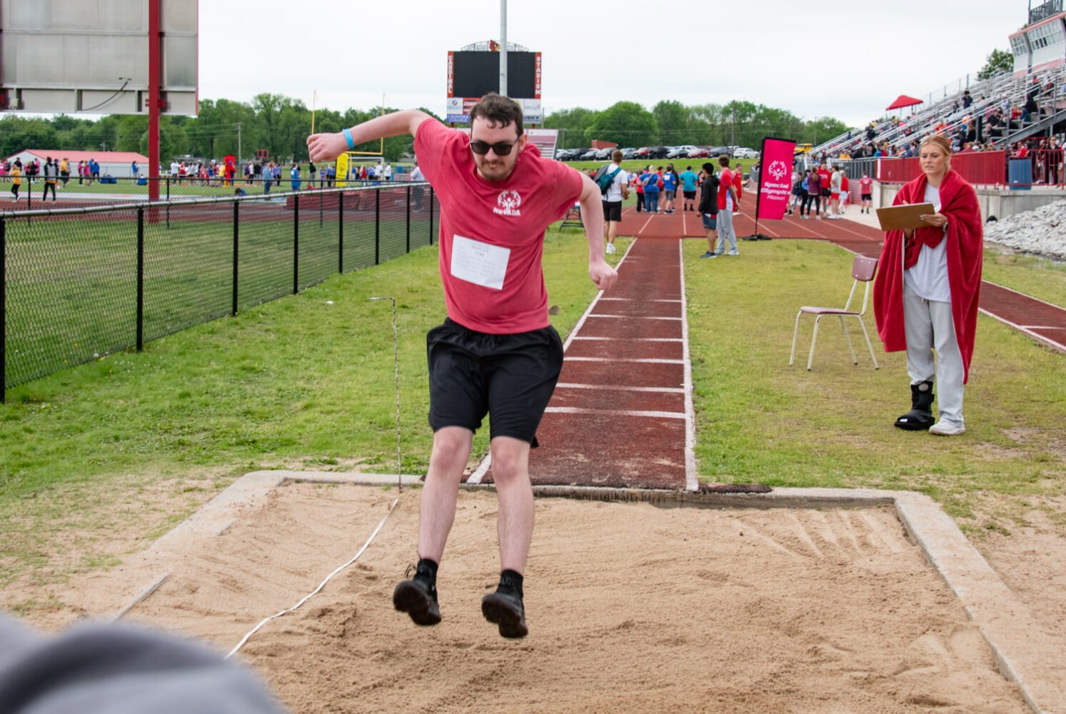 Male athlete in a red shirt and black shorts leaps into a sand pit during a track and field long jump event, with a track and stands in the background.