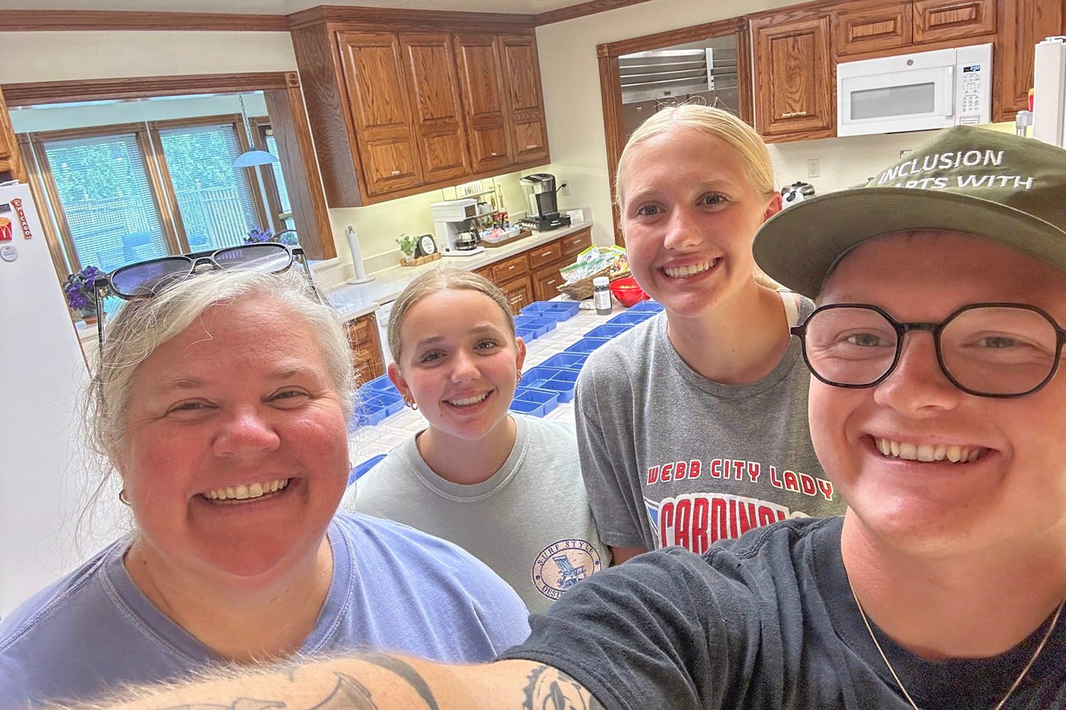 Four smiling people posing for a selfie in a home kitchen with wooden cabinets and blue storage bins on the counter behind them.