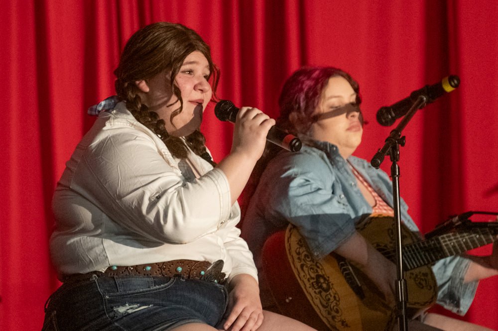 Two female singers perform on a red-curtained stage: one in a white shirt singing into a microphone, the other strumming a decorated acoustic guitar.
