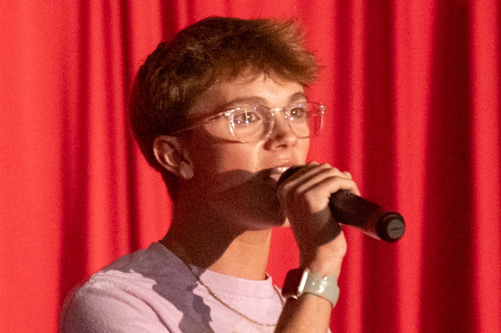 Young person with short hair and glasses speaking into a microphone on a stage with red curtains behind them.