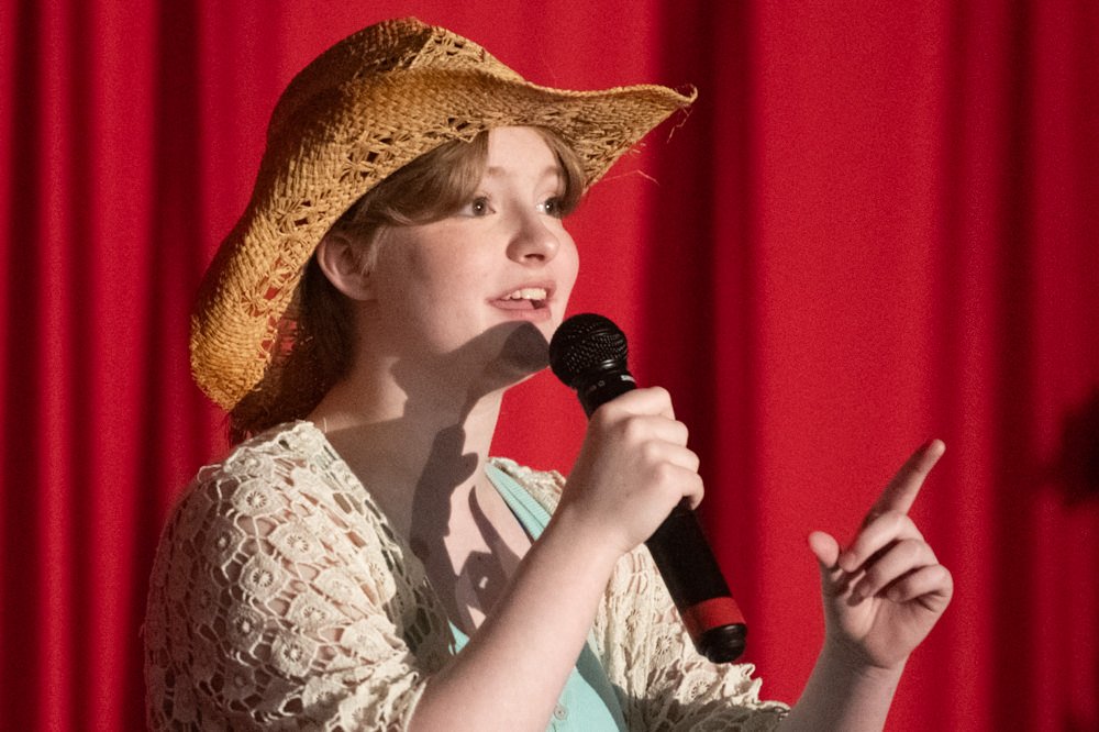 Young girl wearing a straw hat sings into a microphone on a red-curtain stage.