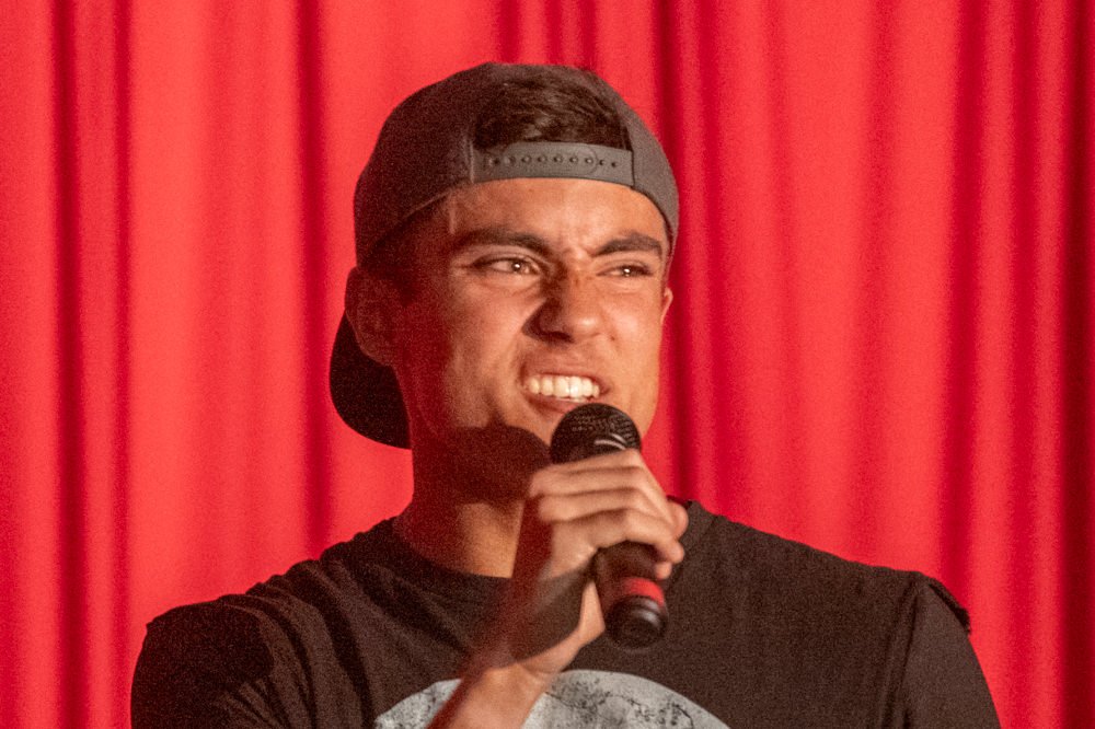 Male performer singing into a handheld microphone on a stage with a red curtain backdrop; wearing a backward baseball cap and black shirt.