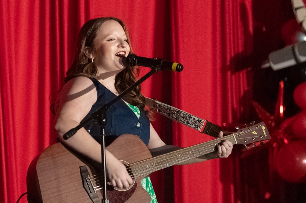 Female singer performing on stage, singing into a microphone while playing an acoustic guitar against red curtain backdrop