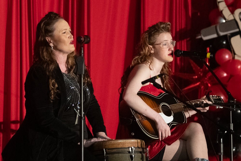 Two female musicians performing on stage: one singing into a microphone while the other plays an acoustic guitar, red curtains in the background.