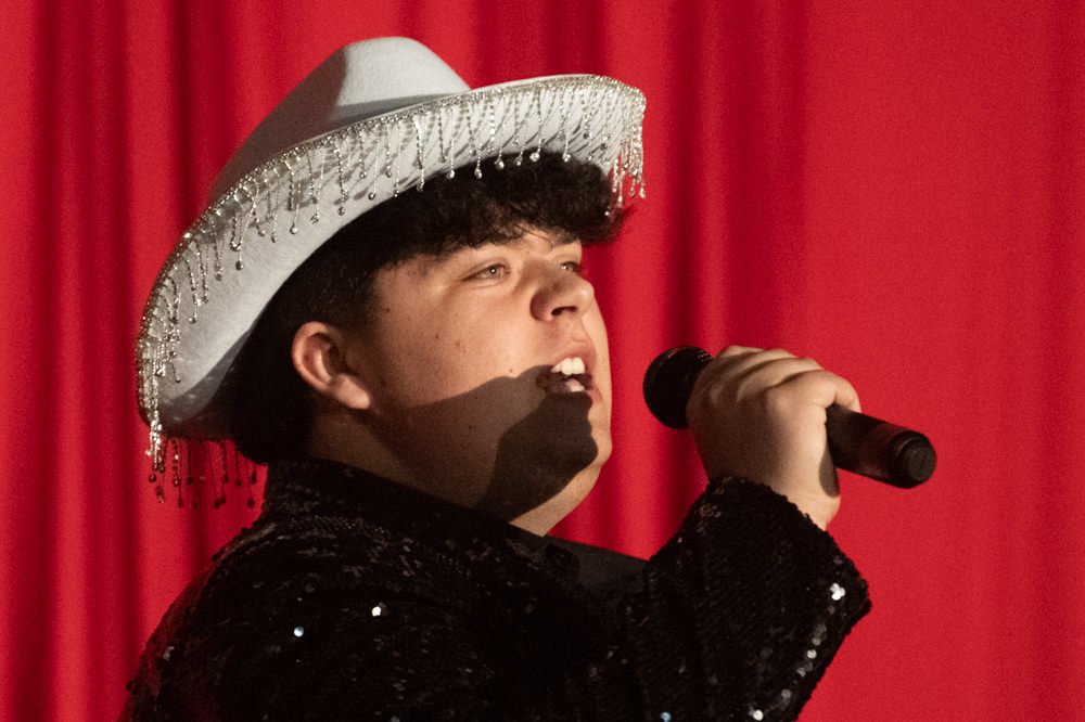 Performer wearing a white fringe-hatted cowboy hat sings into a microphone on a red-stage backdrop of curtains.