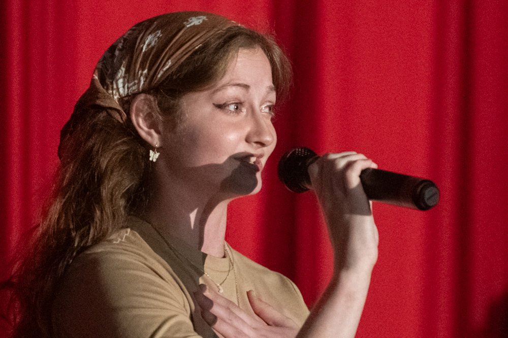 Woman singing into a handheld microphone on stage with red curtains behind her.